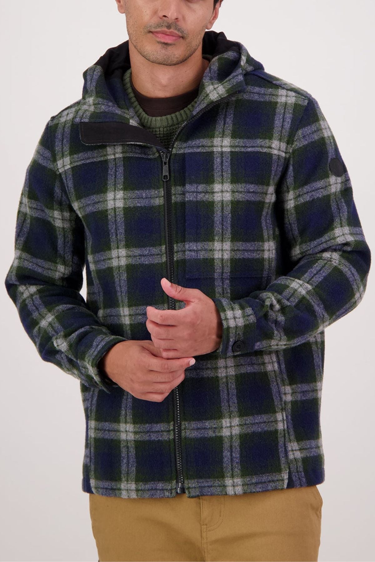 Wearing the Swanndri Hudson Hoodie—a green, blue, and white plaid jacket with a hood—over a green sweater and tan pants, this person highlights the hoodie’s classic style and durable warmth against a plain light background.