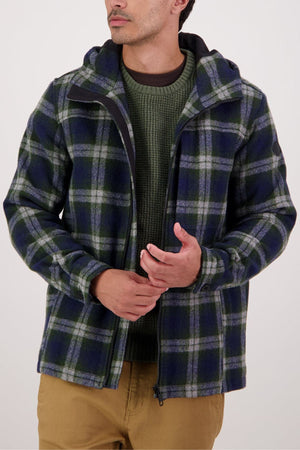 Wearing a green, navy, and gray Swanndri Hudson Hoodie over a green sweater with tan pants, the person stands against a plain light background, head slightly turned, hands loosely clasped.