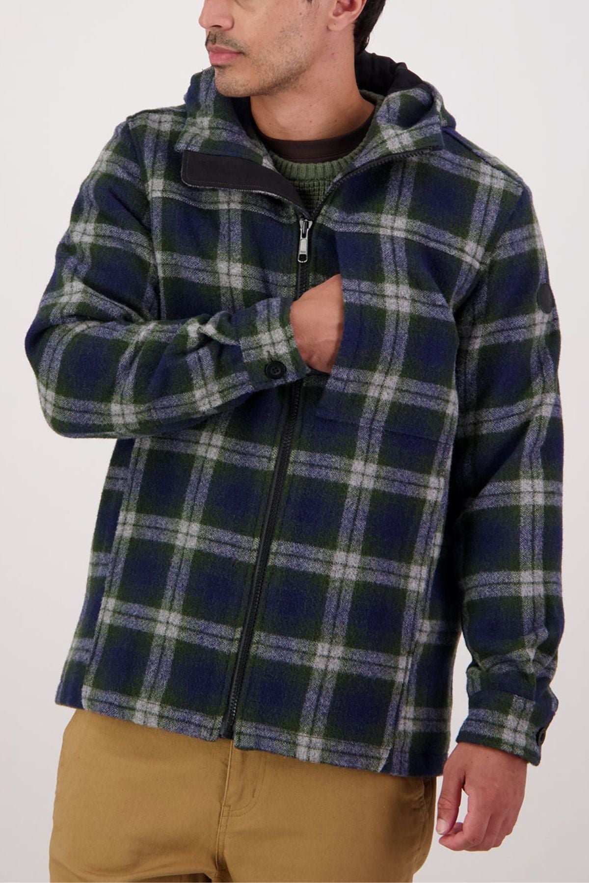 A man in a blue and green plaid Swanndri Hudson Hoodie—a warm, durable jacket made from New Zealand wool—over a green shirt and tan pants is unzipping the front with one hand, his face turned slightly to the side.