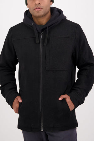 Someone wearing a black Swanndri Hudson Jacket over a black hoodie, hands in jacket pockets, stands against a plain white background.
