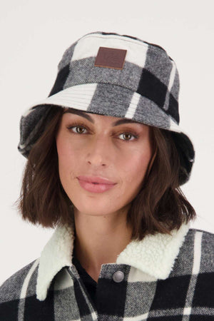 A woman with short brown hair faces the camera with a neutral expression, wearing the Swanndri Kaimai Wool Bucket Hat and a matching plaid jacket with a white sherpa collar.