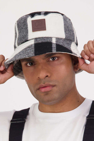 Wearing the Swanndri Kaimai Wool Bucket Hat in black and white, along with a white shirt and black overalls, someone holds the hat's brim with both hands and looks at the camera, ready for any outdoor adventure.