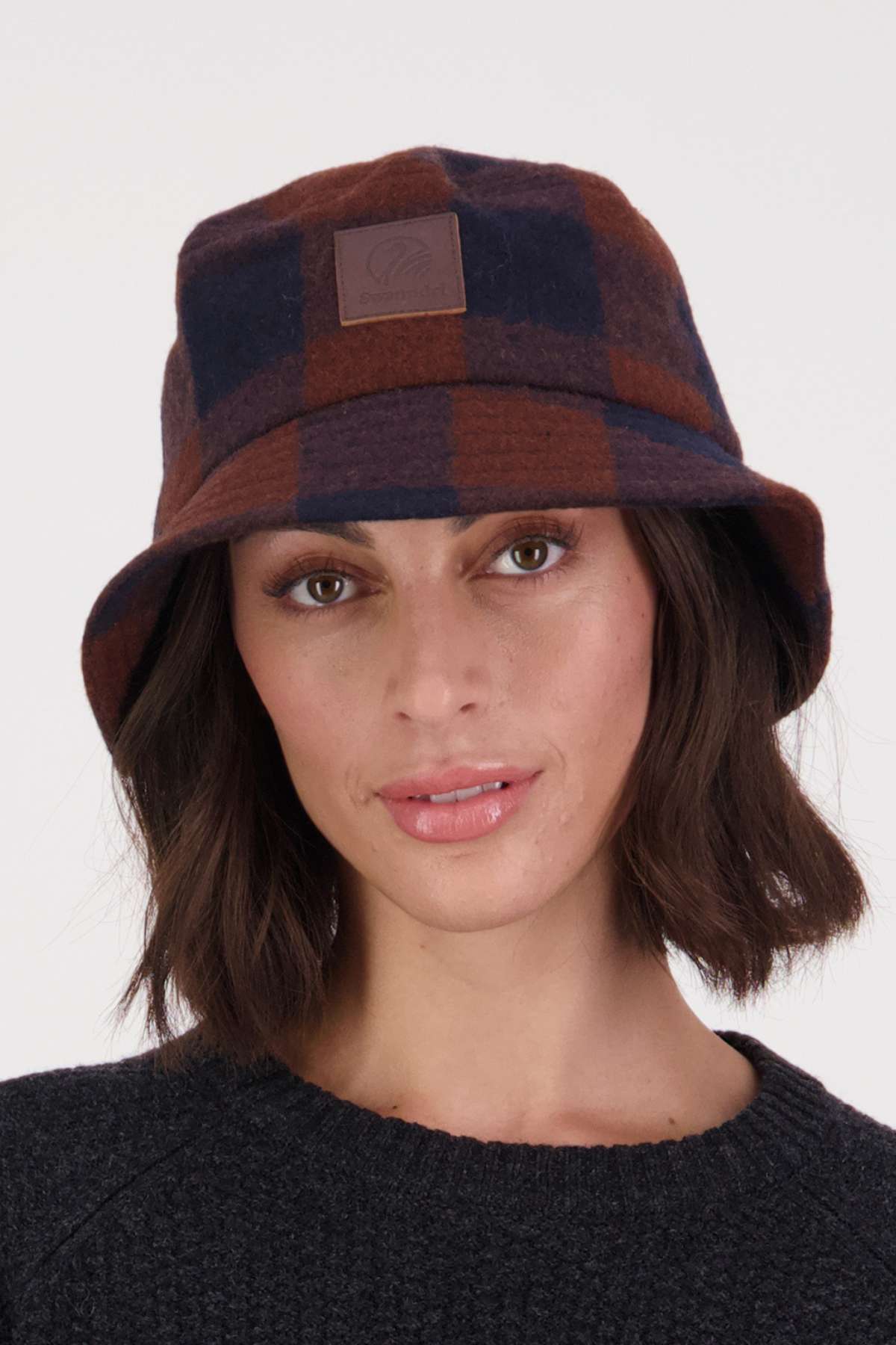 Someone dons a Swanndri Kaimai Wool Bucket Hat in brown and dark blue plaid, paired with a black sweater. They face the camera with a neutral expression, prepared for an outdoor adventure.