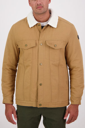 A man wears a Swanndri Kaituna Canvas Jacket with white sherpa lining, buttoned front, and two chest pockets, paired with green pants. The background is light and plain.
