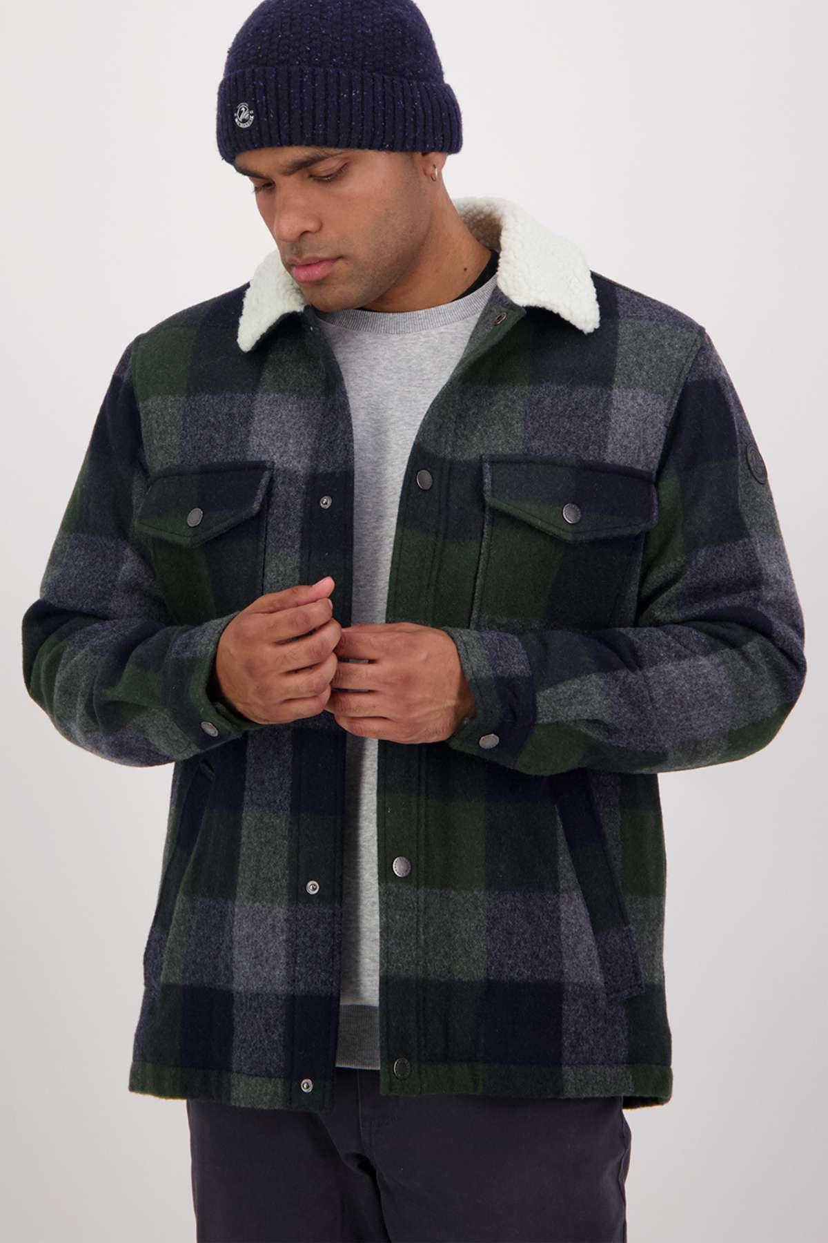 A man in a Swanndri Kaituna Sherpa Lined Jacket featuring a dark green and navy plaid pattern with cozy sherpa lining wears a gray shirt underneath and a navy beanie. Crafted from Merino wool, he adjusts his jacket while looking down against a plain white background.