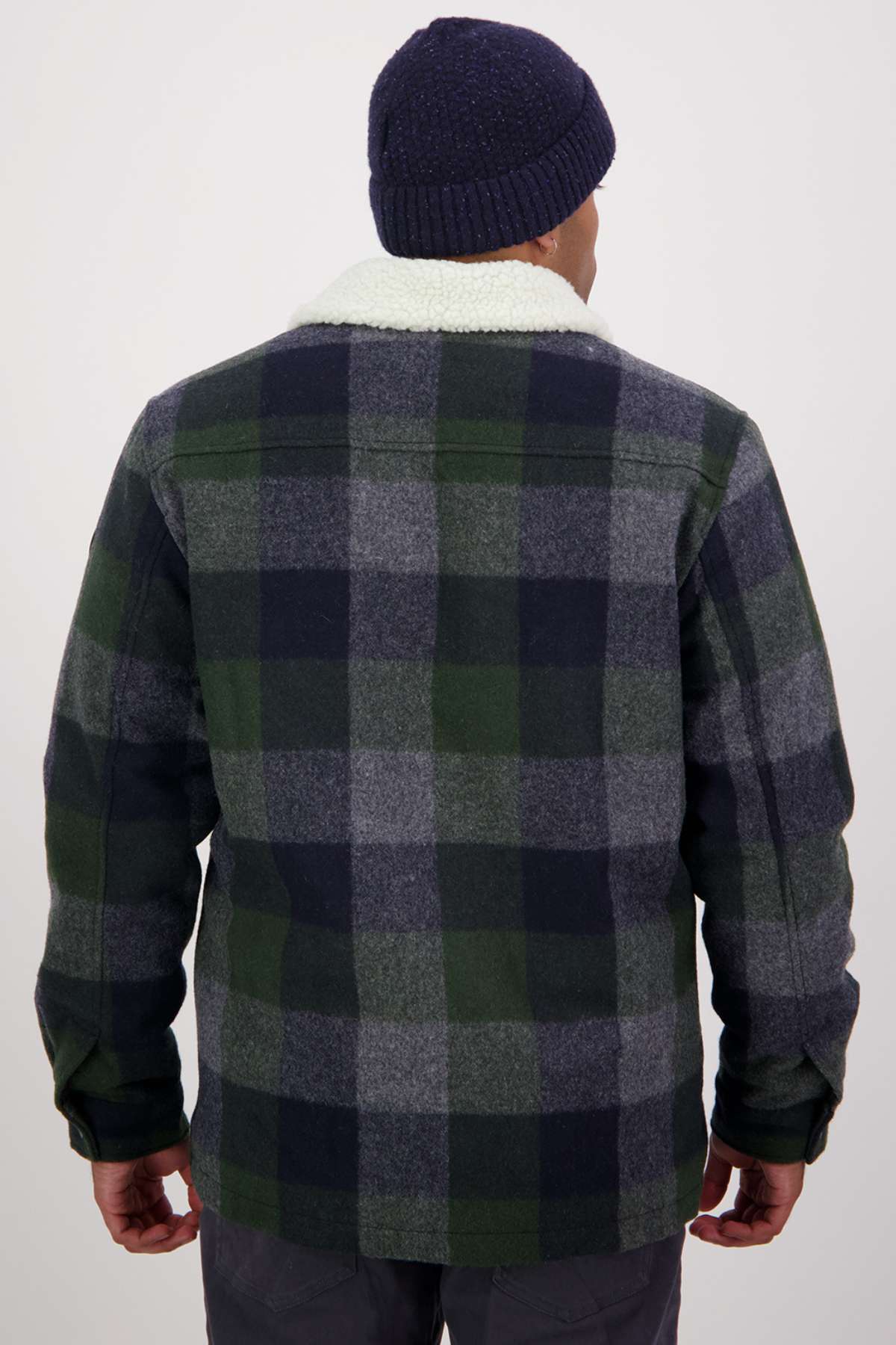 A person is seen from behind in a navy blue beanie and the Swanndri Kaituna Sherpa Lined Jacket, featuring a plaid pattern in green, navy, and gray hues. The jacket's fluffy white sherpa collar adds warmth. The background is plain and light-colored.