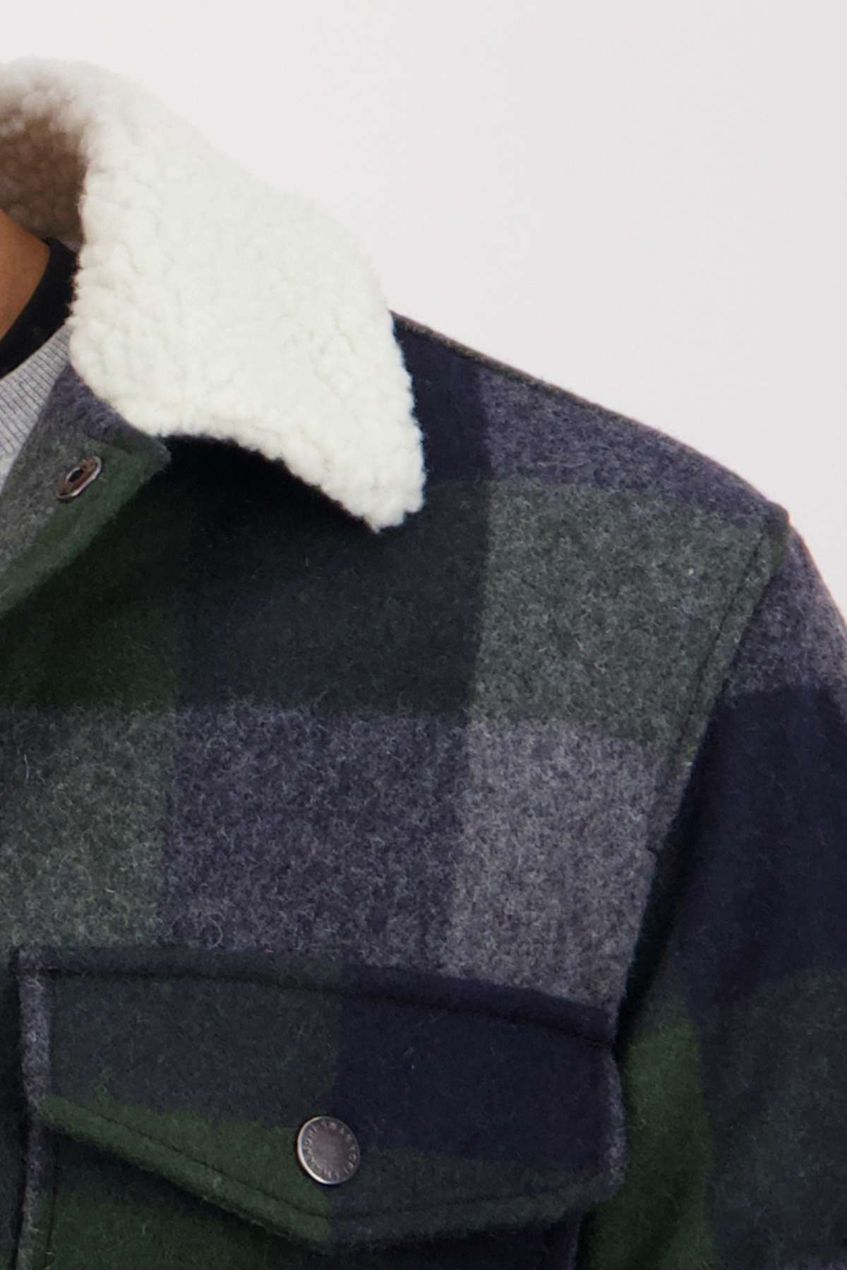 Close-up of the Swanndri Kaituna Sherpa Lined Jacket in green and gray plaid merino wool, showcasing its cozy white sherpa collar and lining, with a metal button and flap pocket on the chest.