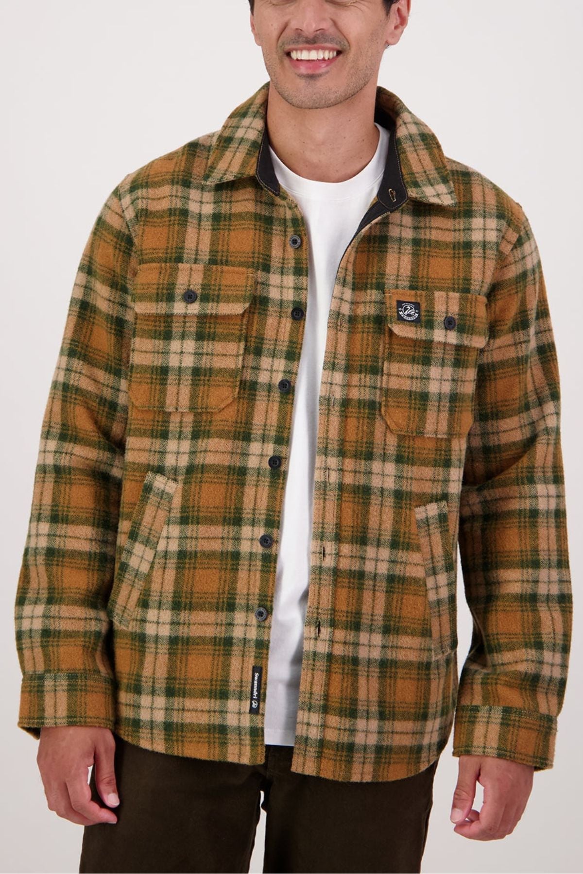 A man wears the Swanndri Kiaraki V4 Wool Shacketโa tan, green, and brown plaid flannel with buttons and chest pocketsโover a white tee and dark pants. He smiles against a plain background.