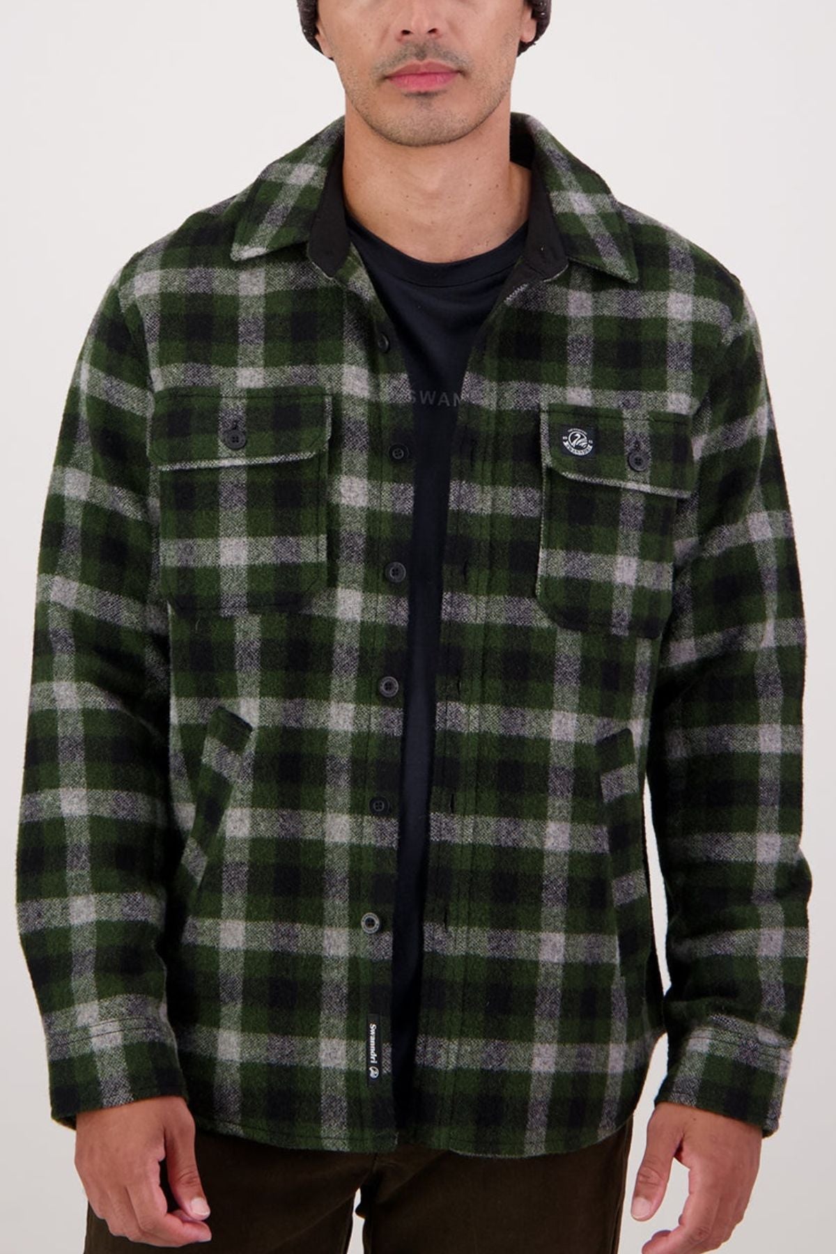 A man wearing a Swanndri Kiaraki V4 Wool Shacket in black and green plaid over a black t-shirt stands with relaxed hands by his sides against a plain light background.