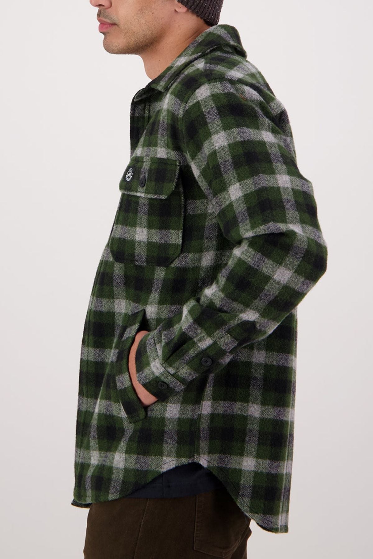 A person stands in profile with one hand in their pocket, wearing the Swanndri Kiaraki V4 Wool Shacket—a green, black, and gray plaid button-up made from New Zealand wool with chest pockets and buttoned flaps. The background is plain and light.