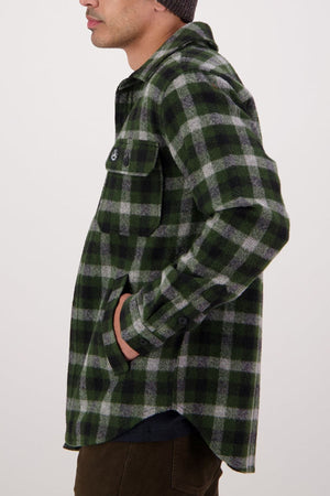 A person stands in profile with one hand in their pocket, wearing the Swanndri Kiaraki V4 Wool Shacket—a green, black, and gray plaid button-up made from New Zealand wool with chest pockets and buttoned flaps. The background is plain and light.