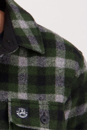 Close-up of the Swanndri Kiaraki V4 Wool Shacket in green and black plaid, featuring a front chest pocket, black button, small round "WAXANDER" label, and visible collar over a dark undershirt.