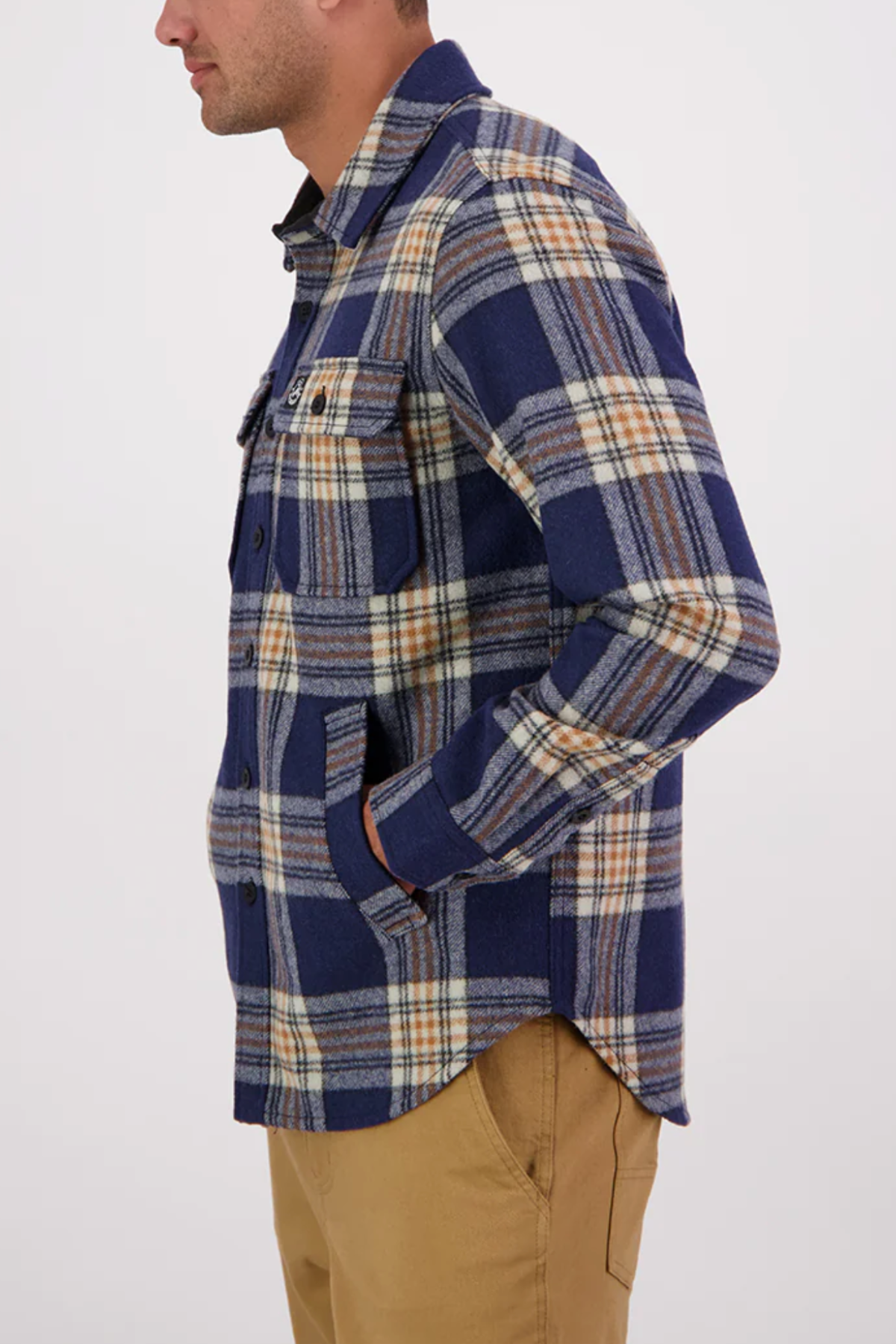 A man in profile wears the Swanndri Kiaraki v3 Wool Shacket—blue, beige, and white plaid with chest pockets—and tan pants. He faces left with his left hand in his pocket against a plain light background.