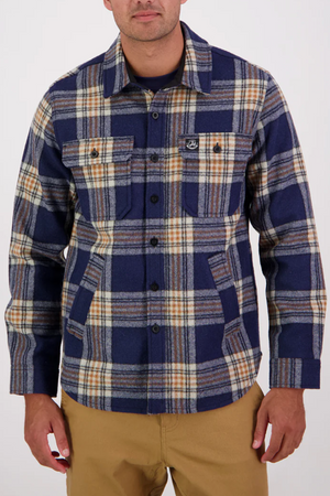 A man wearing the Swanndri Kiaraki v3 Wool Shacket in blue, beige, and white plaid with front pockets and tan pants, made from premium ZQRX wool, stands against a plain light background.