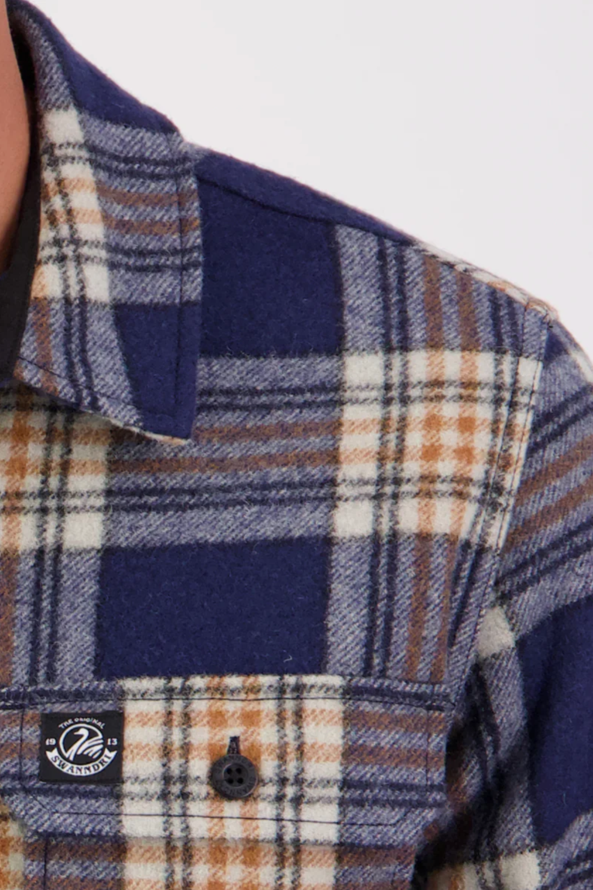 Close-up of the upper left section of the Swanndri Kiaraki v3 Wool Shacket, showing its blue, brown, and white plaid flannel pattern, buttoned chest pocket, and a small black-and-white brand logo on the pocket.