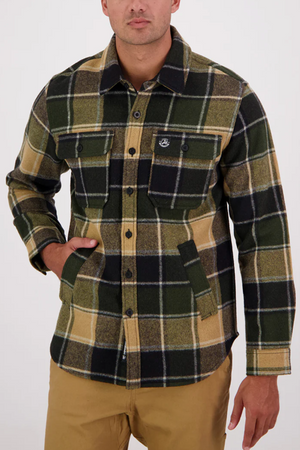 A man wearing a Swanndri Kiaraki v3 Wool Shacket in green, black, and tan plaid with two chest pockets and khaki pants stands against a plain light background, one hand in his pocket.