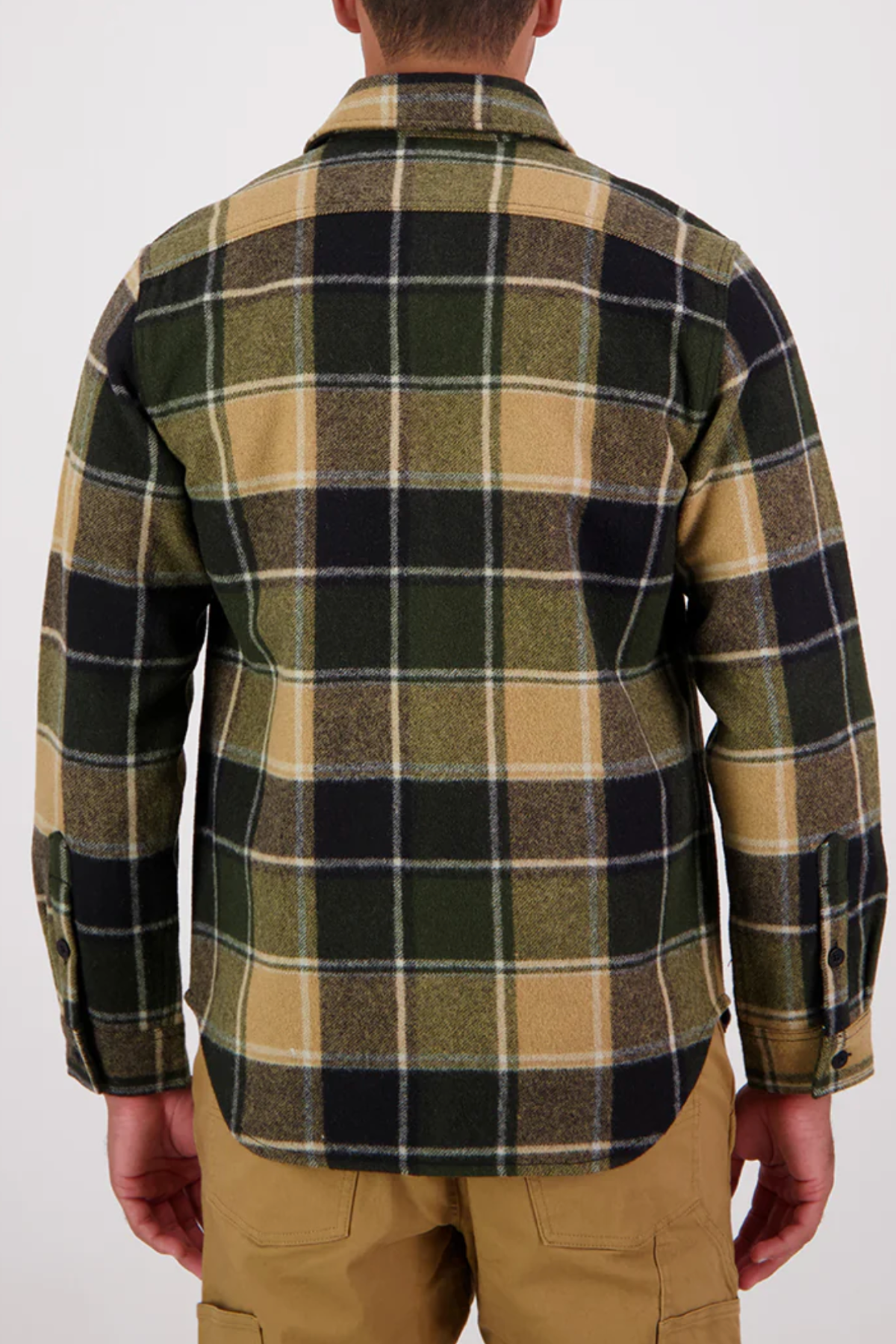A person is seen from the back wearing the Swanndri Kiaraki v3 Wool Shacket in green, black, and beige with long sleeves, paired with tan pants. The background is plain and light-colored.