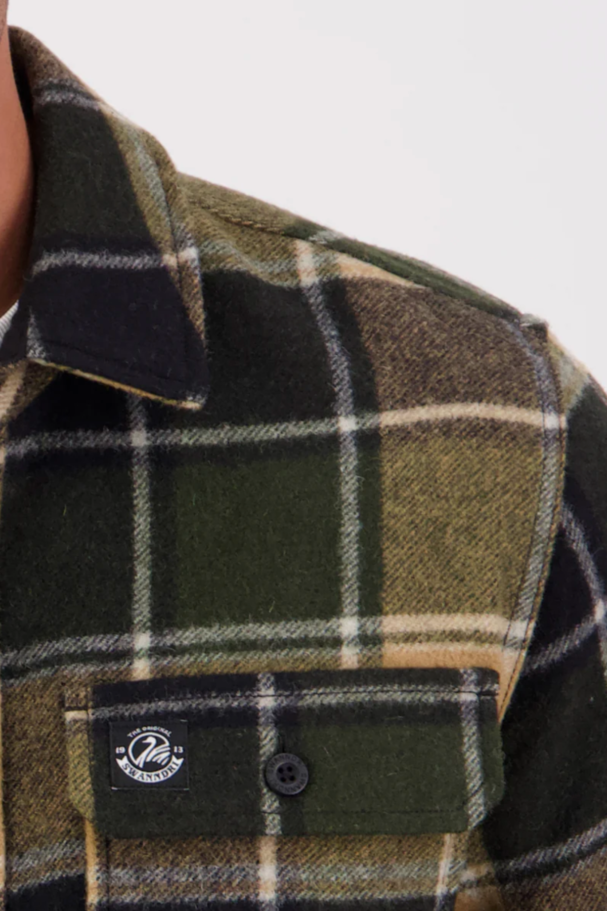 Close-up of the Swanndri Kiaraki v3 Wool Shacket in green, black, and beige plaid. Features a buttoned left chest pocket with a small round logo tag on the flap. The image focuses on the upper shoulder and chest area.