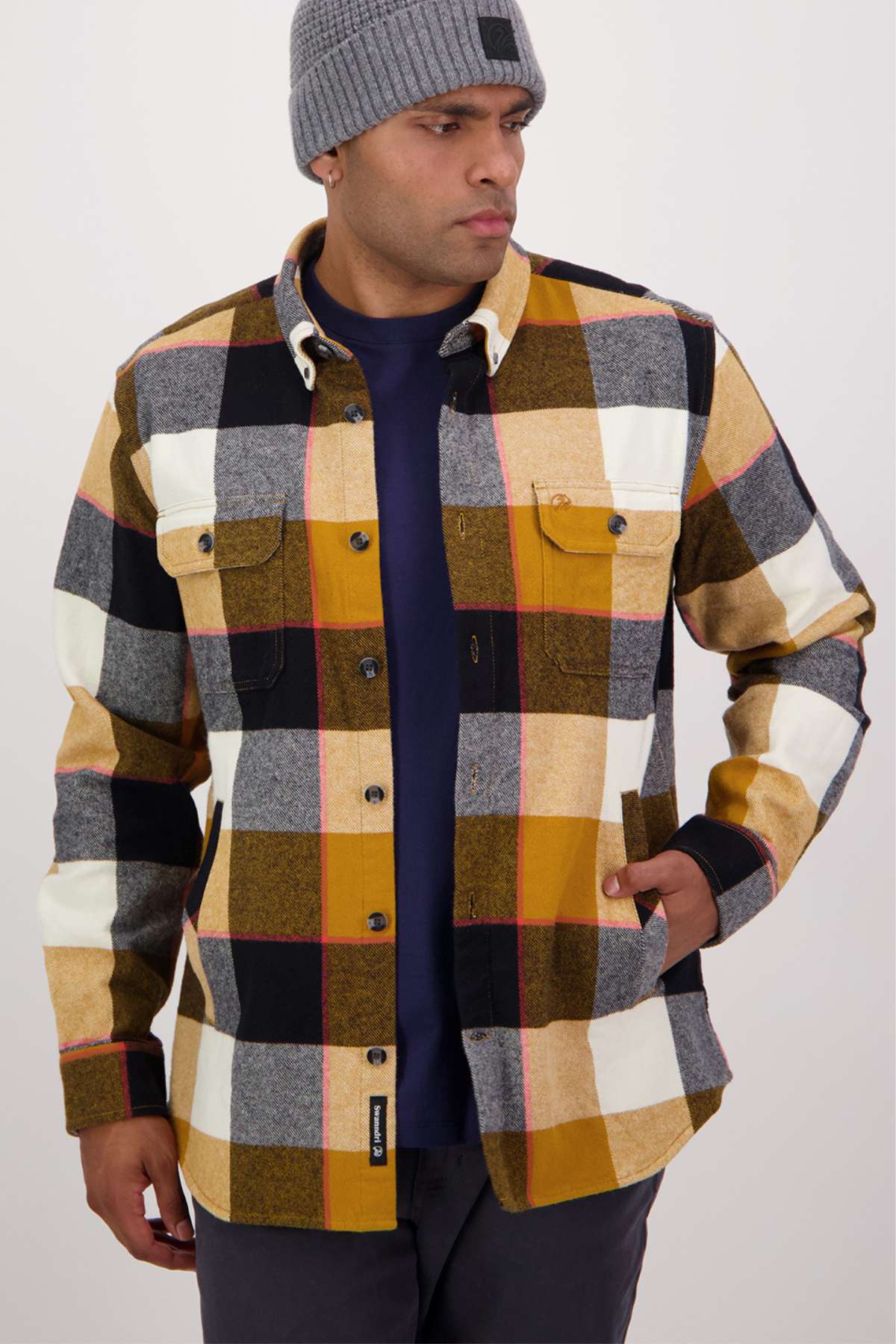 A man in a gray classic fit beanie and a multicolored plaid Swanndri Lowcliffe Shacket made from 100% cotton stands against a plain background, with his navy shirt subtly merging into the scene.