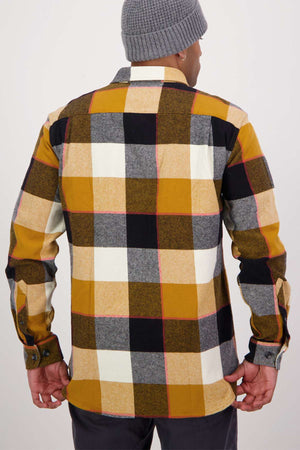 In a classic fit, checkered flannel shirt reminiscent of the Swanndri Lowcliffe Shacket, featuring 100% cotton in mustard, white, black, and gray shades. They pair it with dark pants and a gray beanie as they stand facing away from the camera.