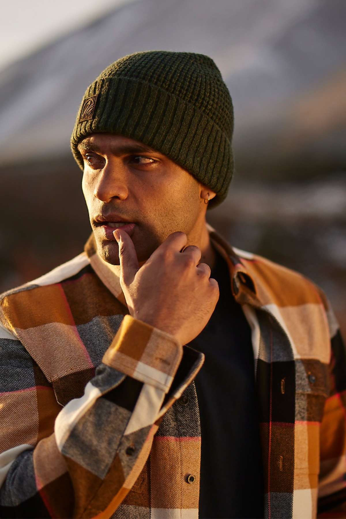 A man in a green knit beanie and a Swanndri Lowcliffe Shacket, appreciated for its classic fit and 100% cotton comfort, stands outdoors. He gazes thoughtfully to the side with his hand near his mouth against a blurred, potentially mountainous backdrop.