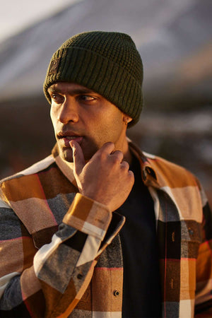 A man in a green knit beanie and a Swanndri Lowcliffe Shacket, appreciated for its classic fit and 100% cotton comfort, stands outdoors. He gazes thoughtfully to the side with his hand near his mouth against a blurred, potentially mountainous backdrop.