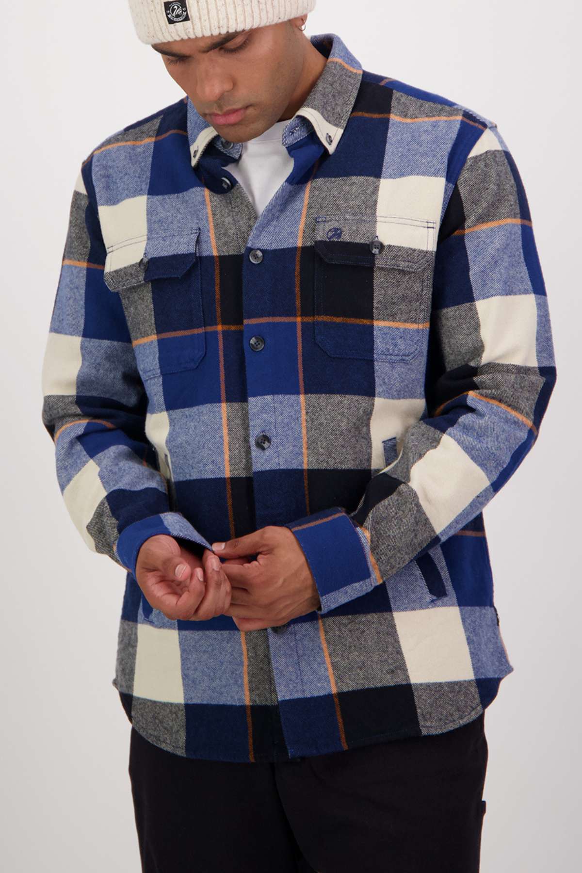 Wearing a Swanndri Lowcliffe Shacket in classic fit blue, black, gray, and white plaid 100% cotton with a white knit beanie, a person looks down touching their hands. The background is plain and light-colored.