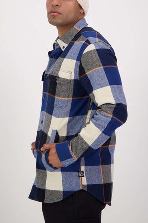 Wearing the Swanndri Lowcliffe Shacket in blue, grey, white, and black plaid with black pants and a white knit beanie, a person stands hands-in-pockets against a plain background.