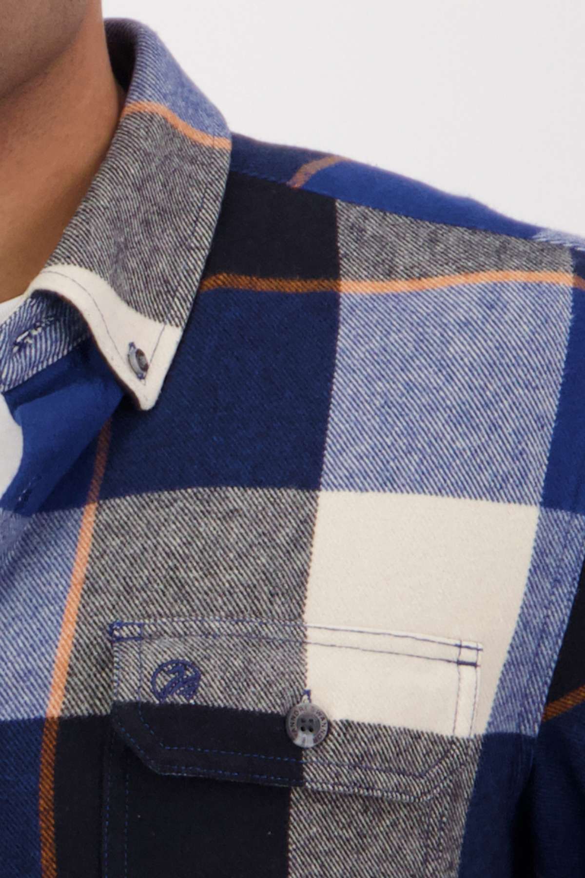 A close-up shows a person wearing the Swanndri Lowcliffe Shacket in blue, black, white, and gray plaid with a classic fit, front pocket, and buttoned collar. Only the lower face, collar, and upper chest are visible.