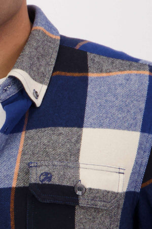 A close-up shows a person wearing the Swanndri Lowcliffe Shacket in blue, black, white, and gray plaid with a classic fit, front pocket, and buttoned collar. Only the lower face, collar, and upper chest are visible.