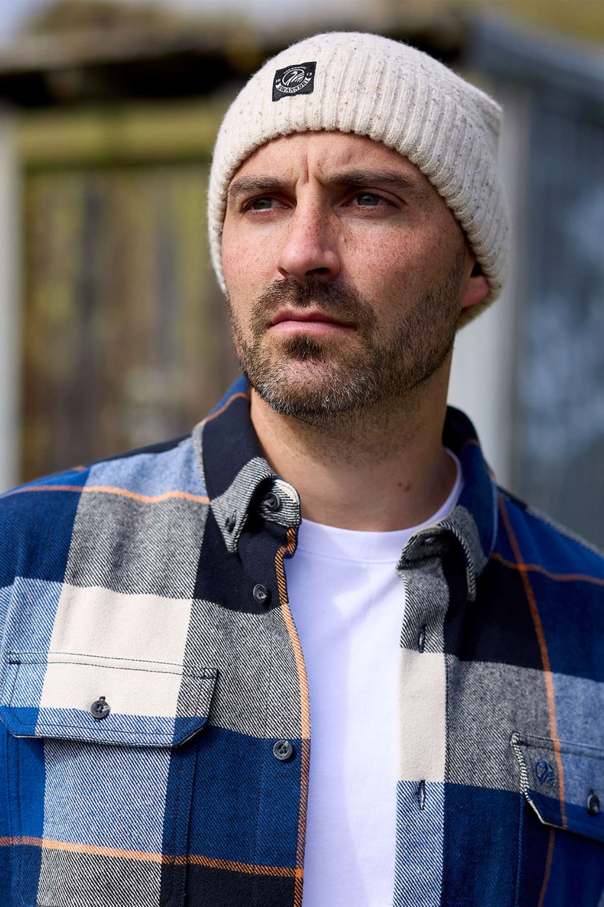 A man with a short beard wears a cream knit beanie and the Swanndri Lowcliffe Shacket in classic fit, featuring blue and gray plaid 100% cotton, layered over a white t-shirt as he gazes into the distance outdoors.