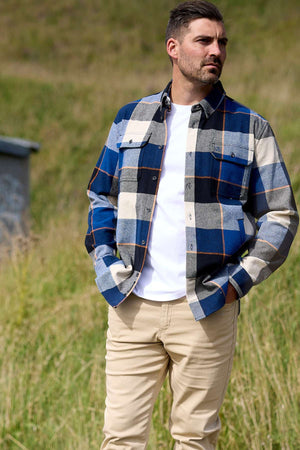 A man with short dark hair and a beard stands outside on grass, wearing a Swanndri Lowcliffe Shacket over a white T-shirt and beige pants, looking to the side with one hand in his pocket.