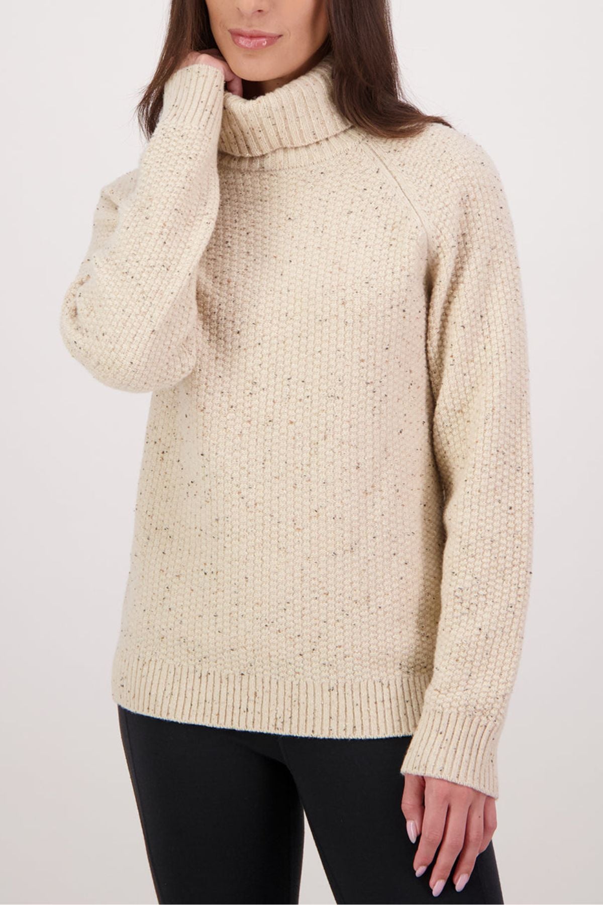 A woman wears the Swanndri Lyttelton v2 Roll Neck Knit, a cream chunky knit sweater with long sleeves, paired with black leggings against a plain light background.