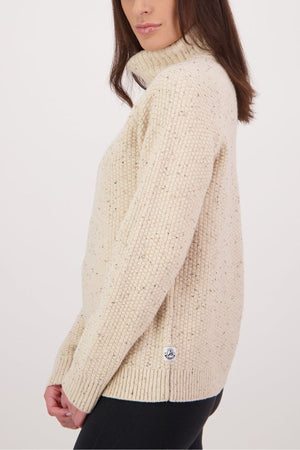 A woman stands sideways against a white background wearing the Swanndri Lyttelton v2 Roll Neck Knit, a cream-colored, chunky lambswool jumper with long sleeves, a subtle speckled pattern, and a small label near the hem.