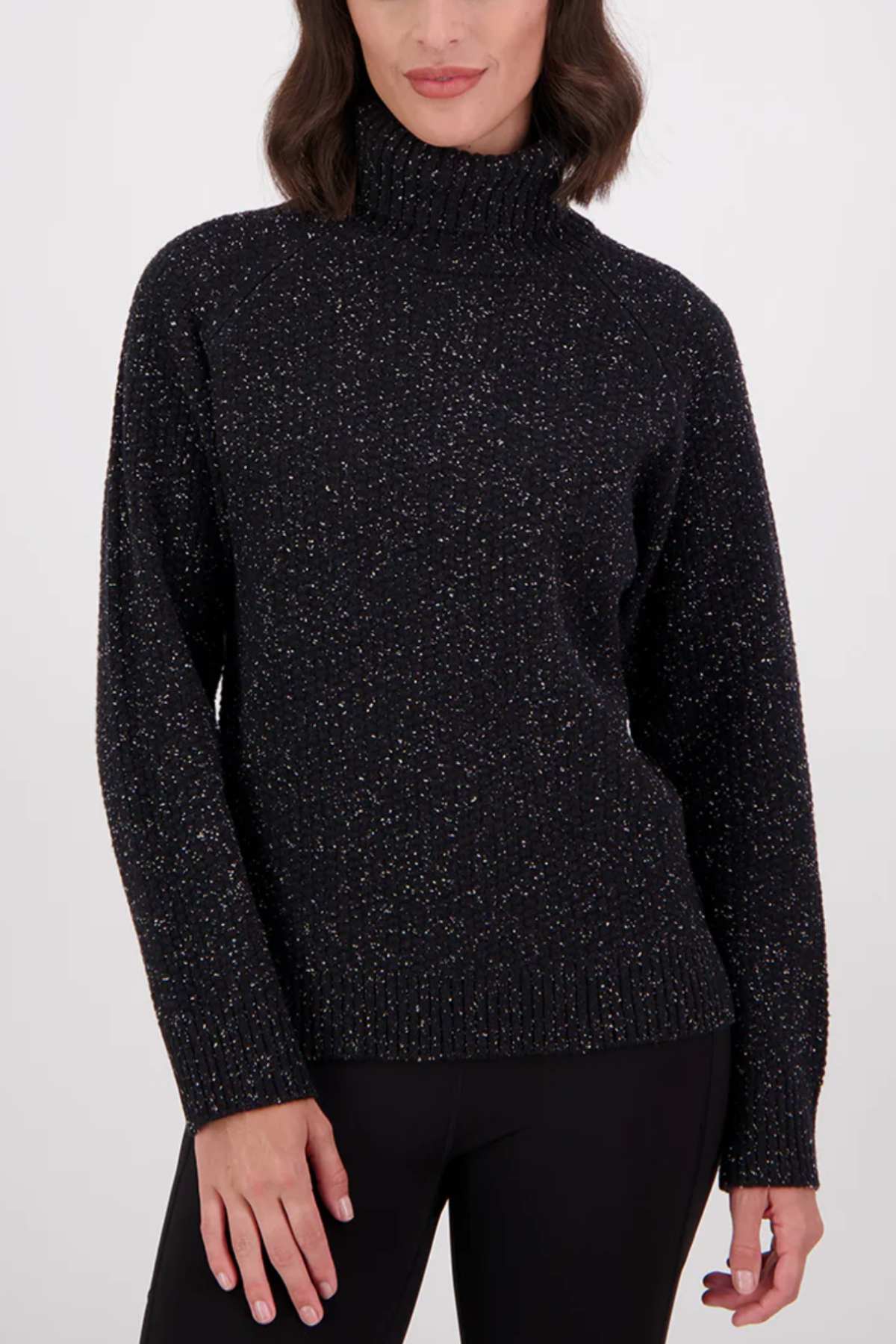 A woman with wavy dark hair wears the Swanndri Lyttelton v2 Roll Neck Knit sweater in black and black pants, standing against a plain light background.