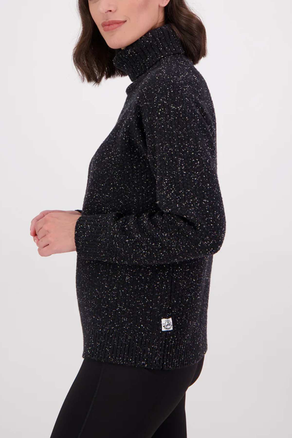 A woman with shoulder-length dark hair wears the Swanndri Lyttelton v2 Roll Neck Knit, a black speckled sweater, paired with black pants. She stands sideways with her left arm slightly bent against a plain white background.