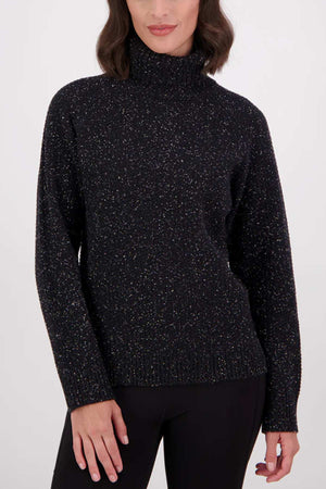 A woman with wavy dark hair wears the Swanndri Lyttelton v2 Roll Neck Knit sweater in black and black pants, standing against a plain light background.