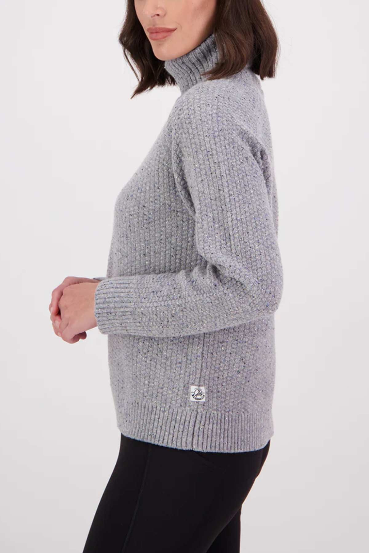 Woman in profile wears the Swanndri Lyttelton v2 Roll Neck Knit in light grey with black pants, standing against a plain white background. She has shoulder-length dark brown hair.