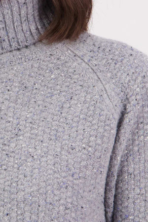 Close-up of the Swanndri Lyttelton v2 Roll Neck Knit in light grey lambswool with speckled texture and ribbed turtleneck, showing only the upper chest, shoulder, and part of the neck and hair.