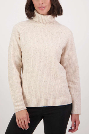 Wearing a Swanndri Lyttelton v2 Roll Neck Knit, a person with shoulder-length dark hair stands against a plain white backdrop. Their cream sweater features a speckled texture, and they pose with one hand tucked slightly into their black pants.