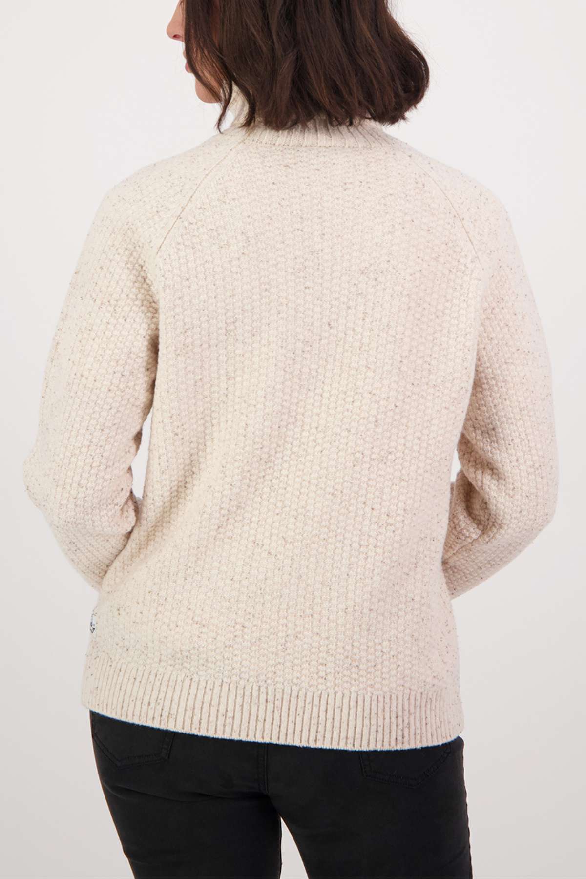 The image depicts someone with dark hair in a cream Swanndri Lyttelton v2 Roll Neck Knit, which features a ribbed collar and hem. They face away from the camera, wearing dark pants against a plain backdrop.