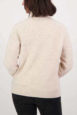 The image depicts someone with dark hair in a cream Swanndri Lyttelton v2 Roll Neck Knit, which features a ribbed collar and hem. They face away from the camera, wearing dark pants against a plain backdrop.
