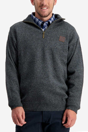 A man is wearing a Swanndri Mariner Zip Neck Sweater in dark gray over a blue and white plaid shirt. The knit sweater features a ribbed hem and cuffs, with a leather patch on the left chest. He is standing against a plain light background.