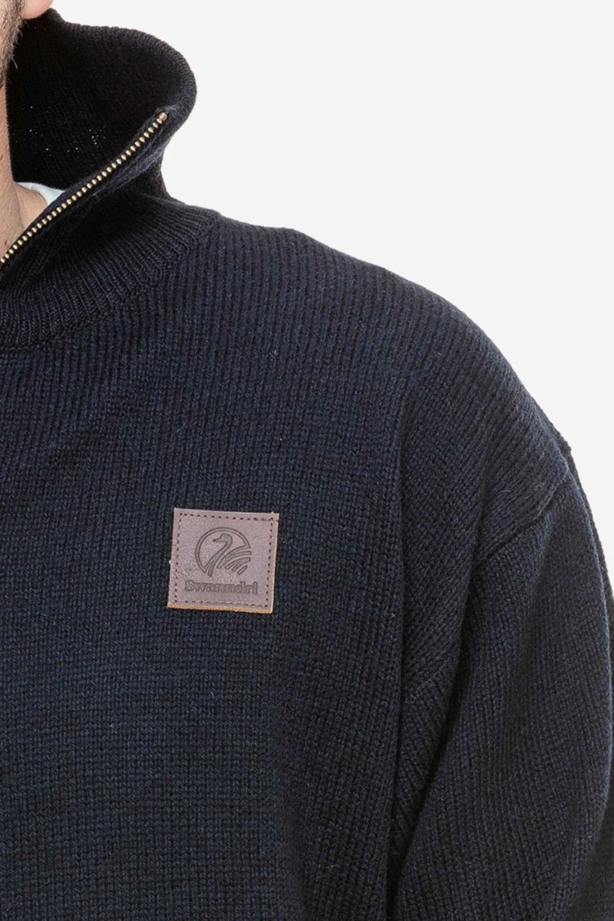 Close-up of a person wearing the Swanndri Mariner Zip Neck Sweater, featuring dark blue ribbed wool with a high collar and half zip. The stylish knit includes a brown leather patch embossed with text and logo on the chest against a plain white background.