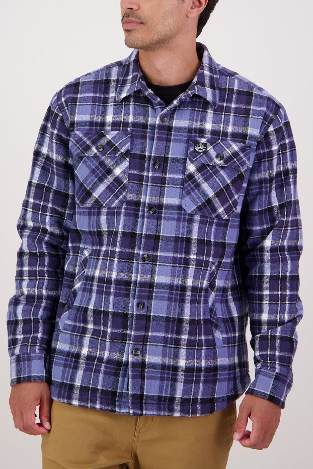A person wearing a purple and blue plaid Swanndri Morven Sherpa Shirt with two chest pockets and a button-up front stands against a plain light background, their head turned slightly to the side.