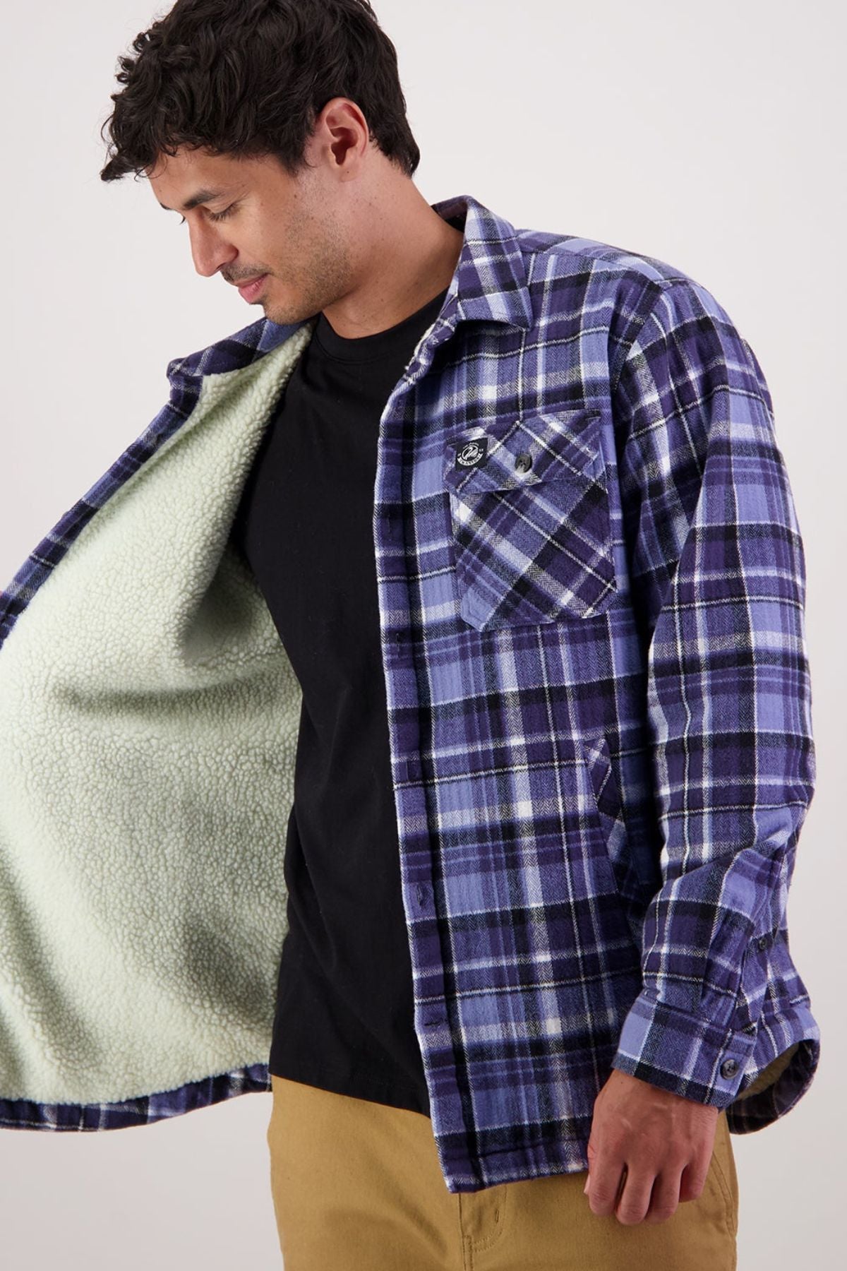 A man in tan pants and a black Swanndri Morven Sherpa Shirt opens a blue/purple plaid jacket to reveal a soft, light green sherpa fleece lining. He looks down and smiles slightly against a plain background.