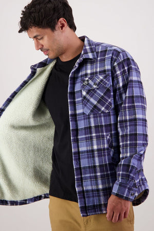 A man in tan pants and a black Swanndri Morven Sherpa Shirt opens a blue/purple plaid jacket to reveal a soft, light green sherpa fleece lining. He looks down and smiles slightly against a plain background.