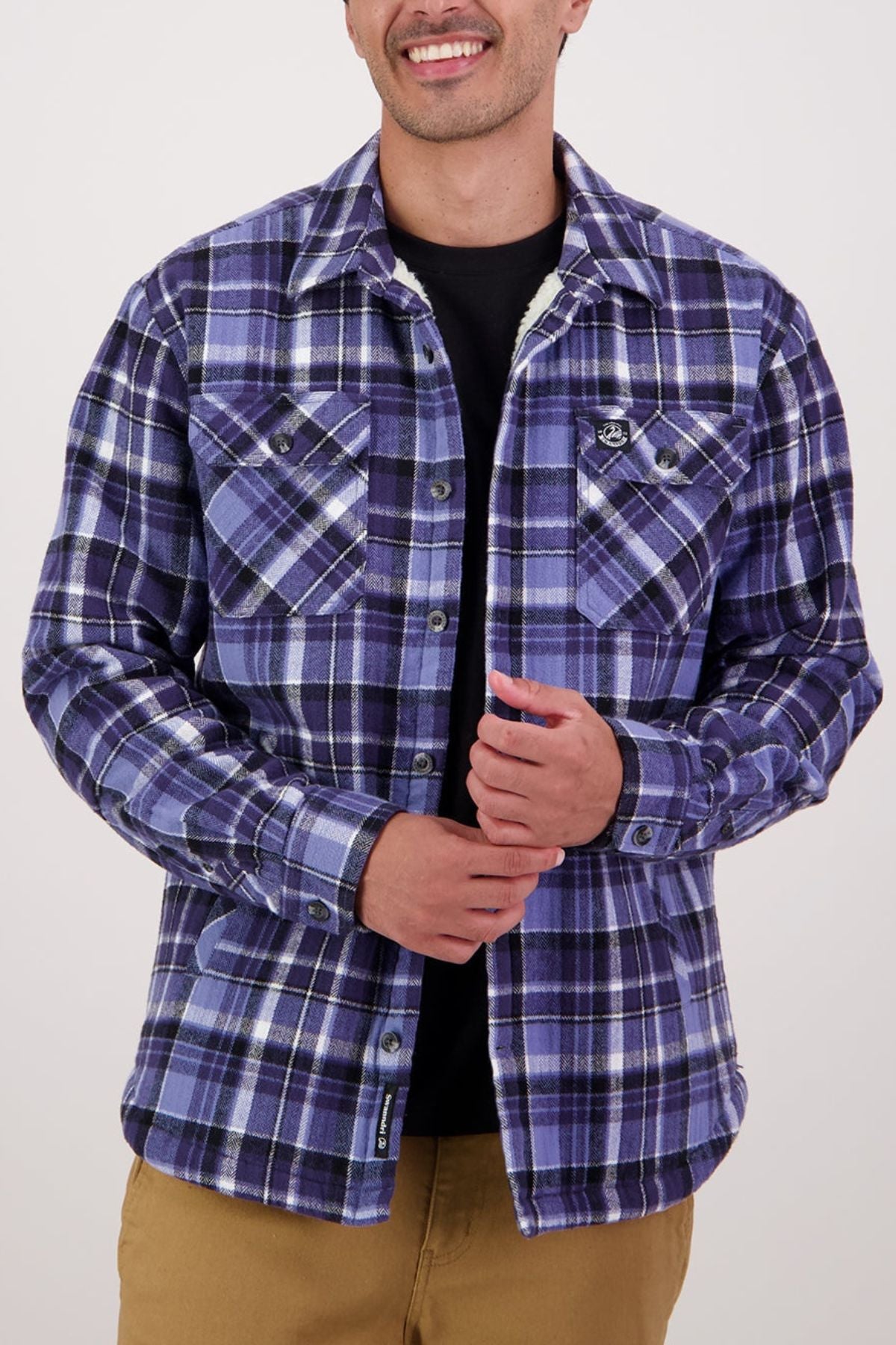 A smiling person wearing a blue and purple Swanndri Morven Sherpa Shirt over a white top and khaki pants, hands held together at chest level.