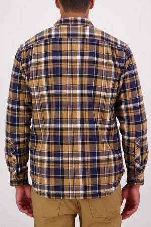 A man is seen from behind in the Swanndri Morven Sherpa Long Sleeve Shirt, featuring brown, navy blue, and white plaid, buttoned cuffs, and tan pants. The 100% cotton shirt is set against a plain light background.