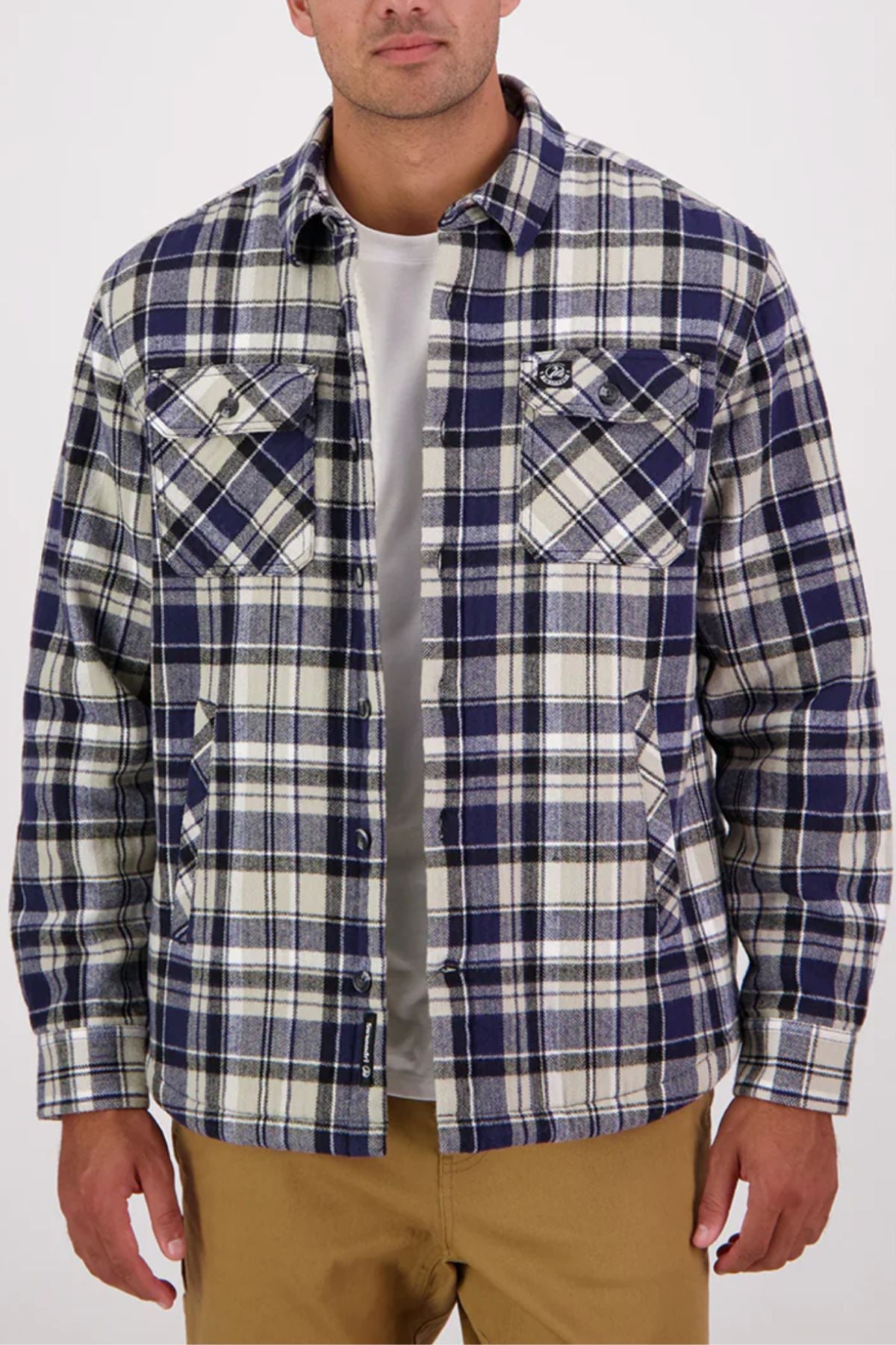 A man wearing a Swanndri Morven Sherpa Long Sleeve Shirtโblue, white, and gray plaid flannel over a white t-shirt and tan pantsโstands against a plain light background.